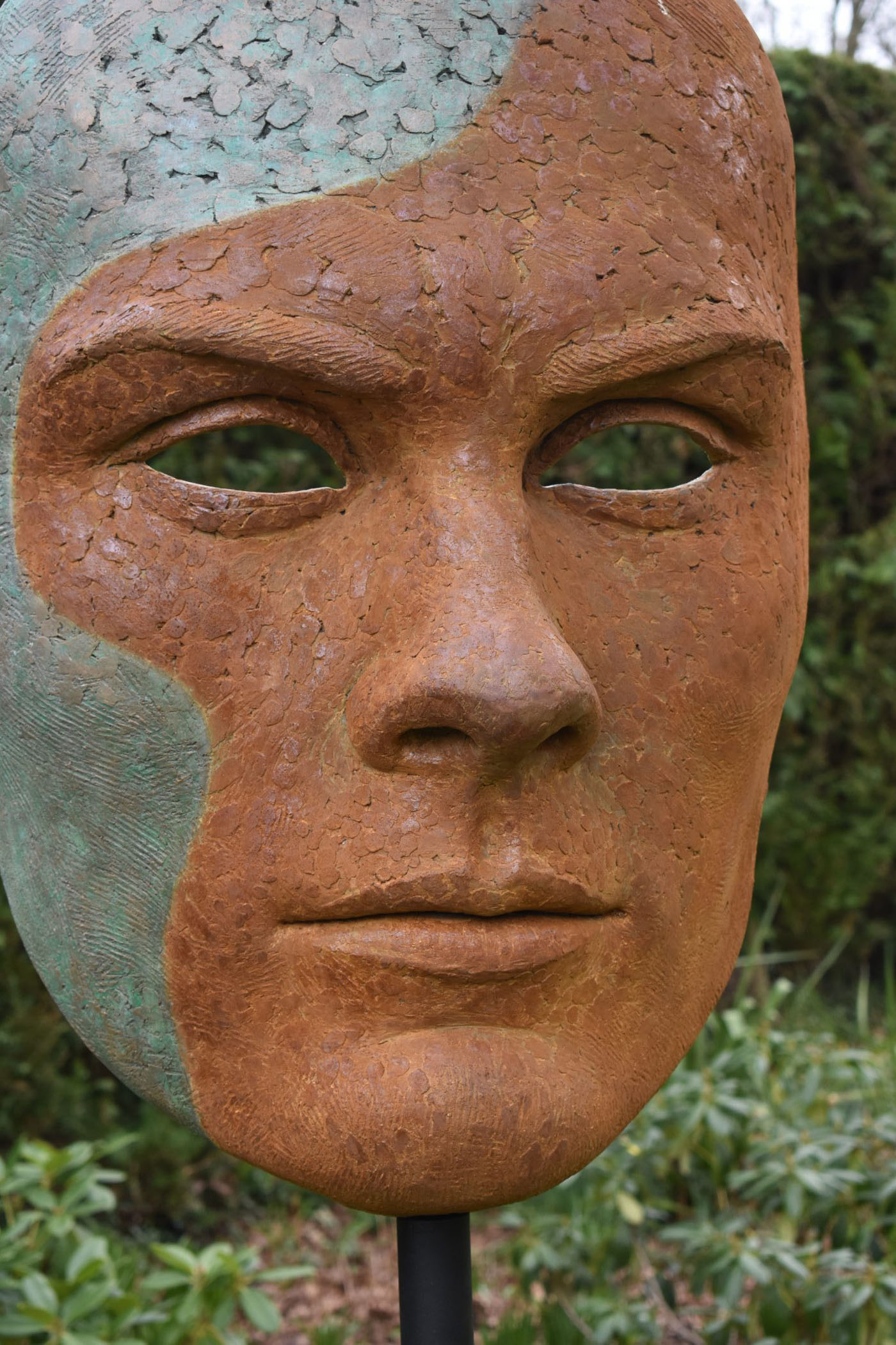 Mask of Achilles by Nicola Godden. A large bronze resin mask sculpture of the face of the Greek warrior, Achilles. Displayed on top of a wooden plinth.