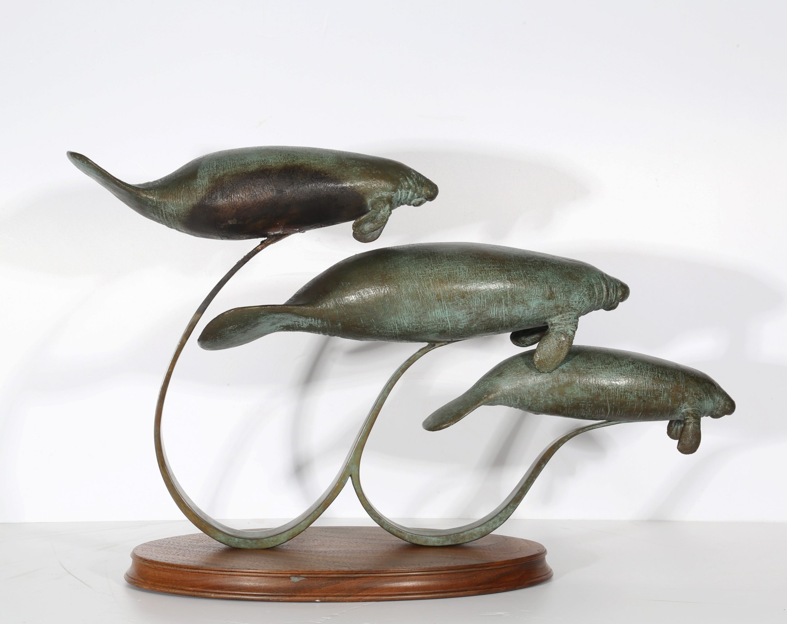 River Shadows by Andre Harvey is a Realistic bronze sculpture of 3 Manatees behind one another, each one slightly higher than the one before it. They are all being held up by a bronze rod to give them the effect of floating/swimming. On a wooden base.
