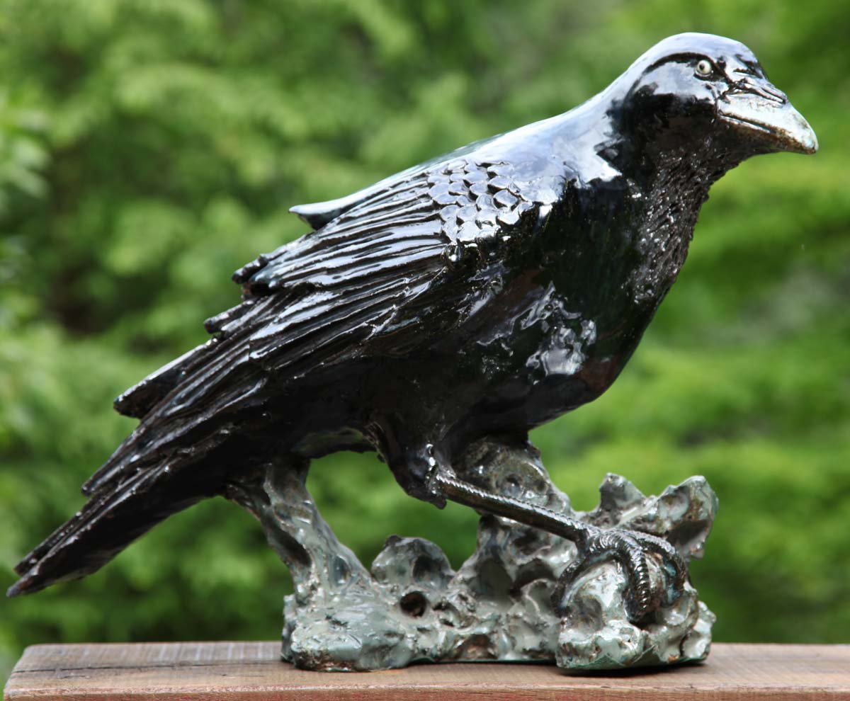 A modern stoneware sculpture of a black raven perched on a stand and looking around.