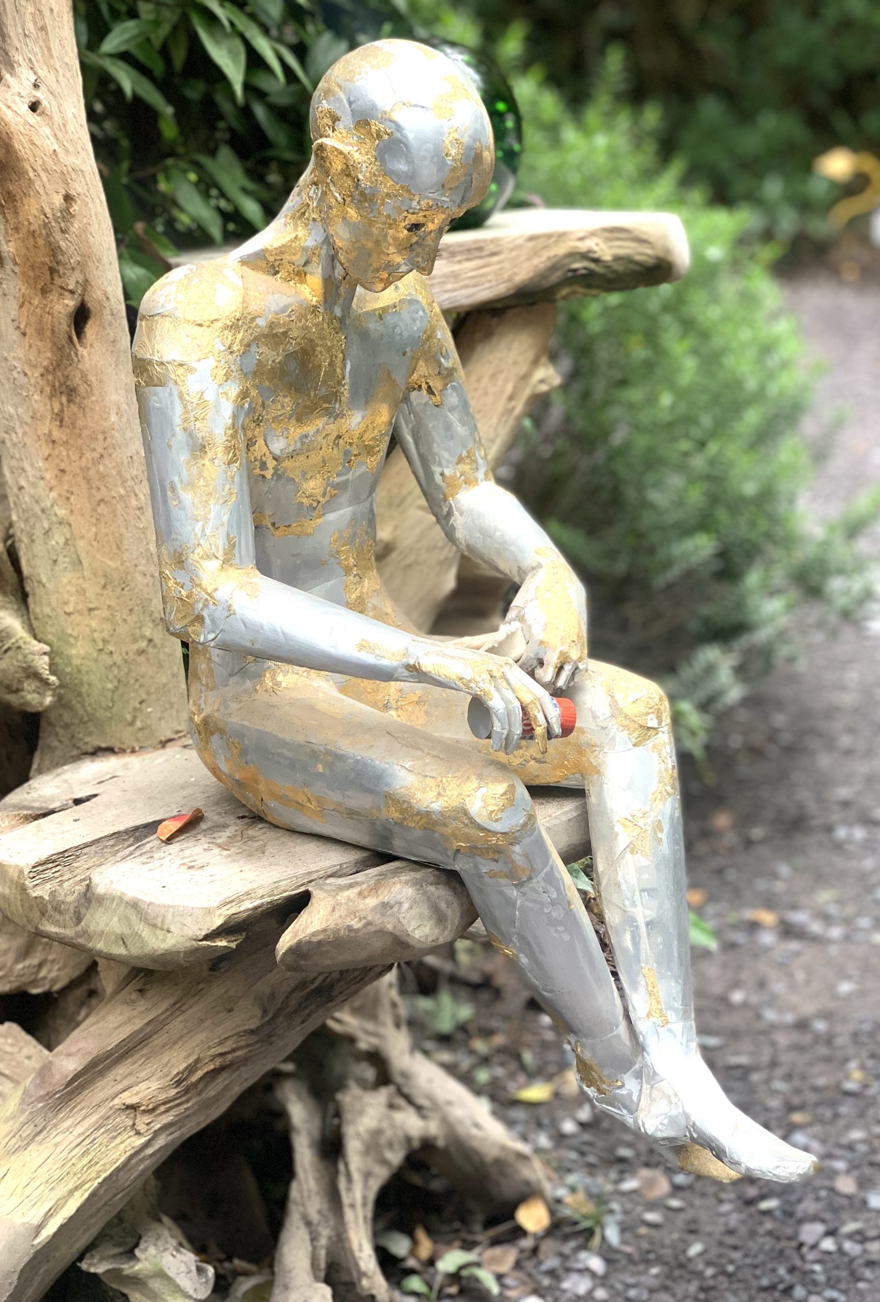 A abstract figurative sculpture of a man sitting while holding two items, made from aluminium.