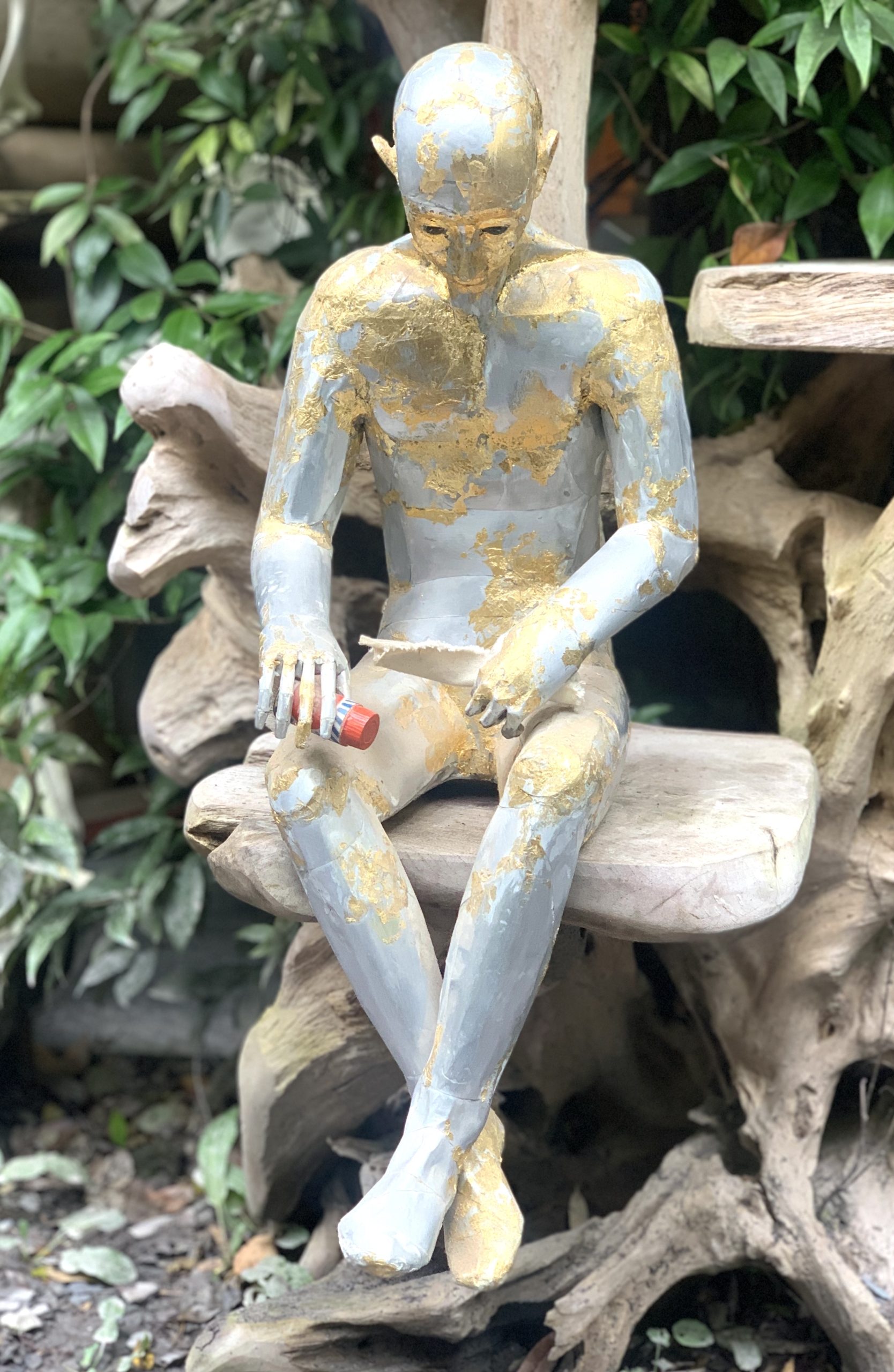 A abstract figurative sculpture of a man sitting while holding two items, made from aluminium.