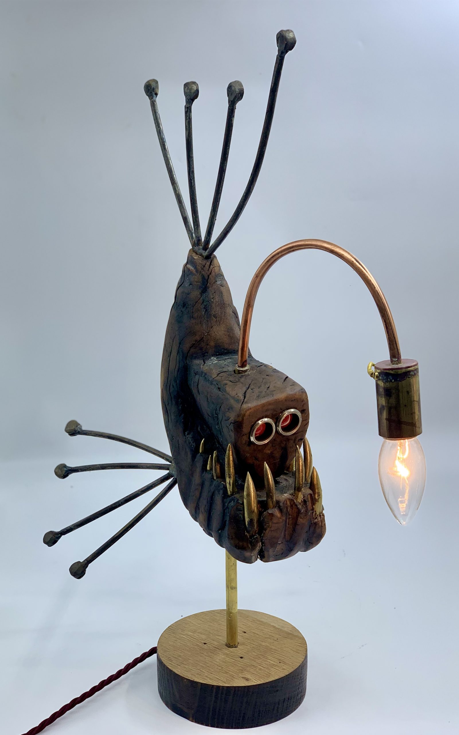 A figurative metal sculpture of a angler fish with a working light, mounted onto a block of wood.