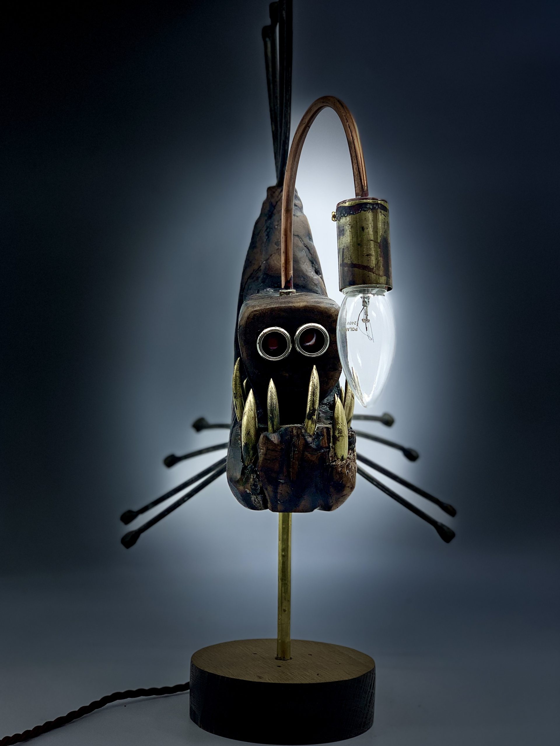 A figurative metal sculpture of a angler fish with a working light, mounted onto a block of wood.
