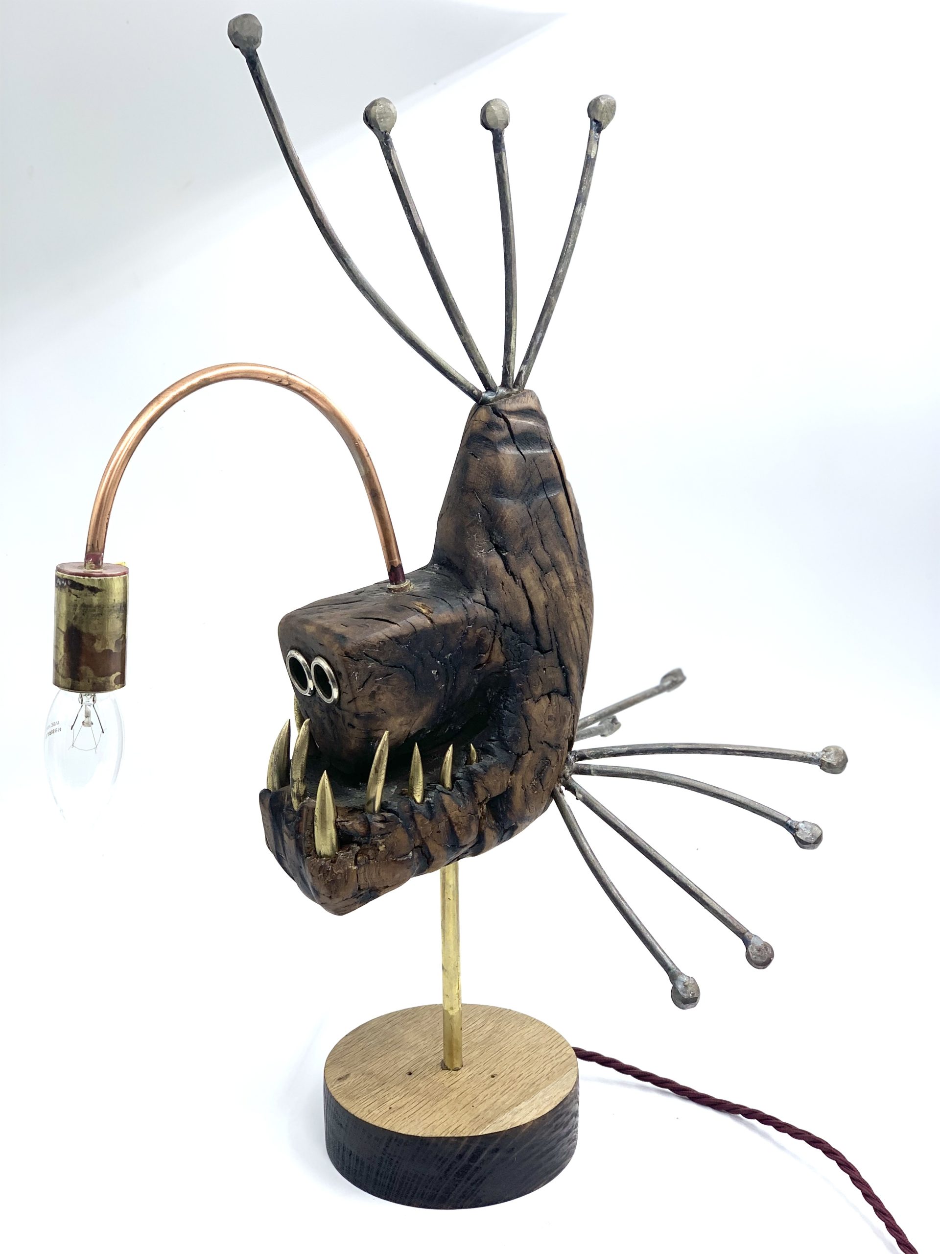 A figurative metal sculpture of a angler fish with a working light, mounted onto a block of wood.