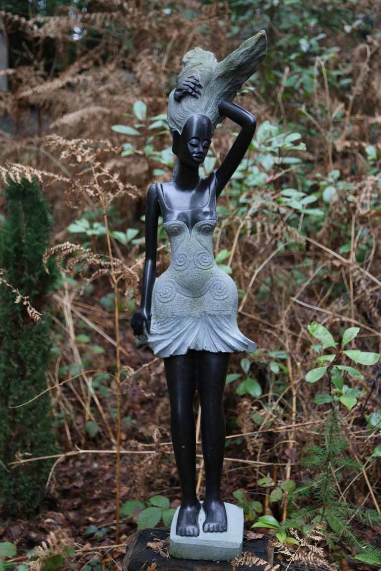 A shona springstone sculpture of a woman standing while holding back her hair.