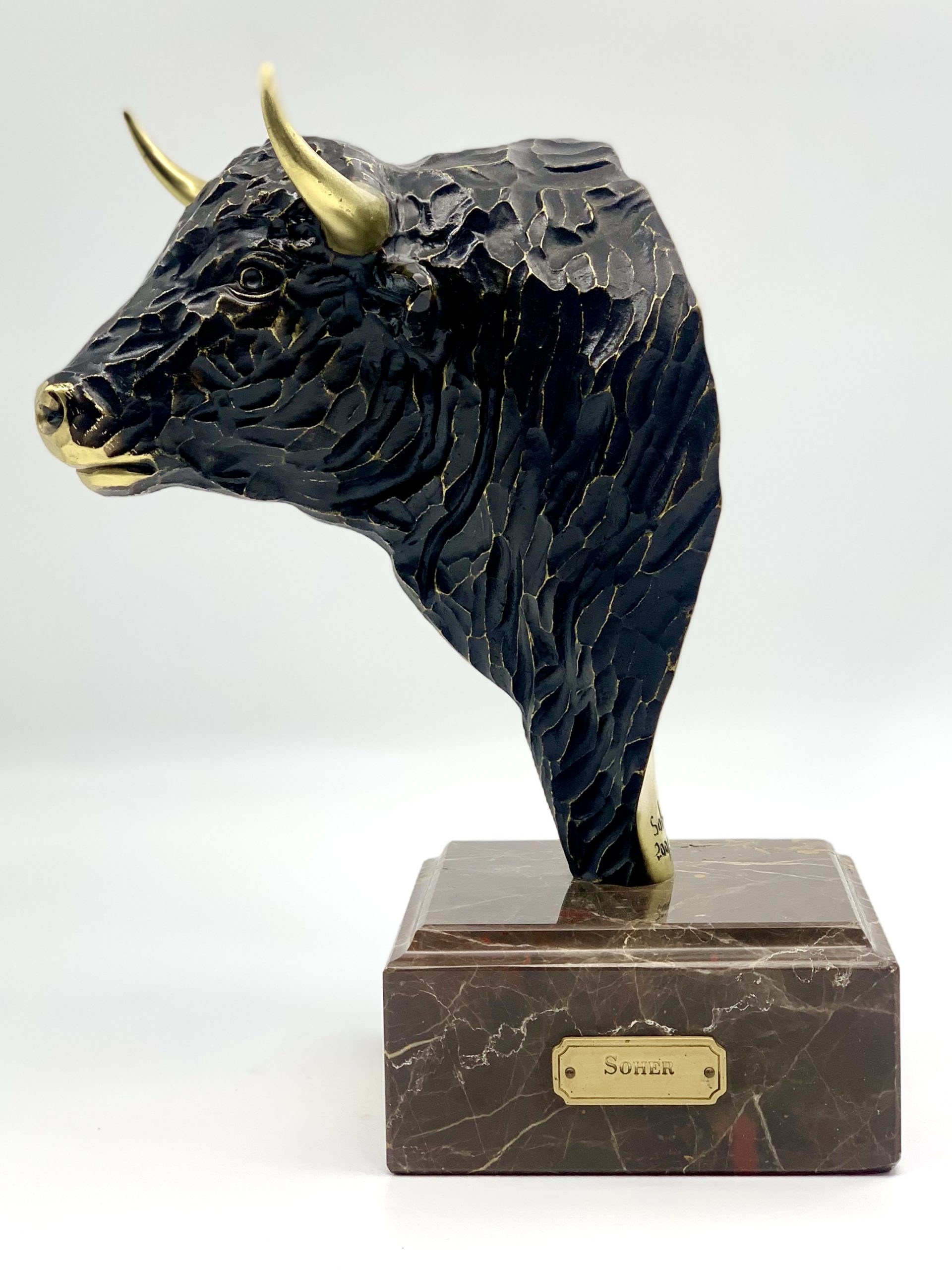 A figurative bronze sculpture of a bulls head with golden horns and nose, mounted onto a block of marble.