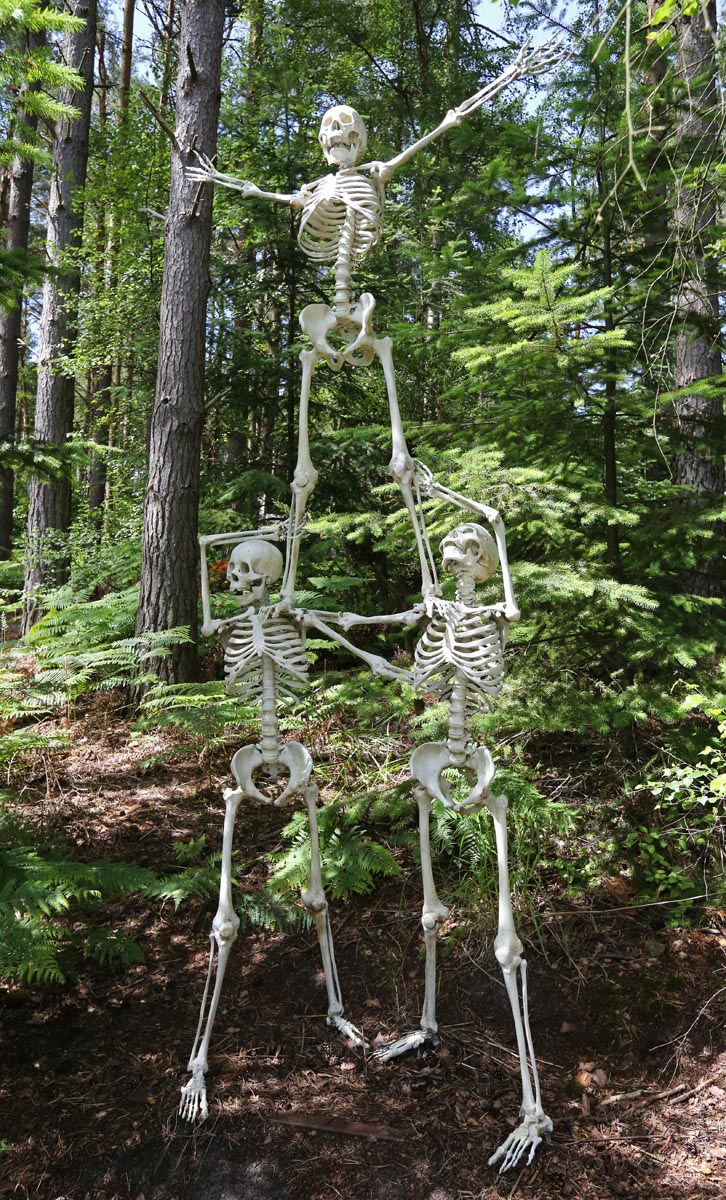 A figurative bronze sculpture of a group of three skeletons with one balancing on top of the other two.
